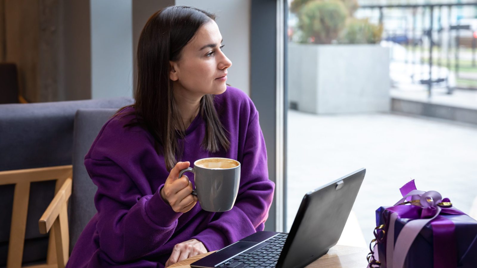 Fiatal nő lila pulóverben egy kávézóban egy csésze kávéval és egy ajándékdobozzal egy laptop előtt.