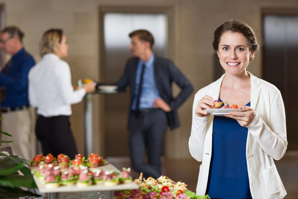 Closeup of smiling at camera beautiful middle-aged woman eating snacks from plate and standing at table with food at buffet reception with blurry people in background